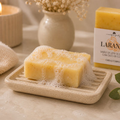 Sabão amarelo com espuma sobre prato cerâmico com embalagem branca e texto em ambiente com vela, vaso e toalha