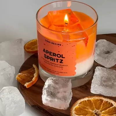 Vela aromática Aperol Spritz em copo de vidro com cubos e fatias de laranja seca.