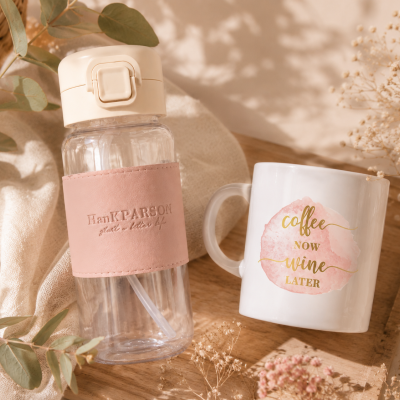 Garrafa de plástico com proteção rosa e caneca branca com texto colorido sobre base de madeira e plantas secas