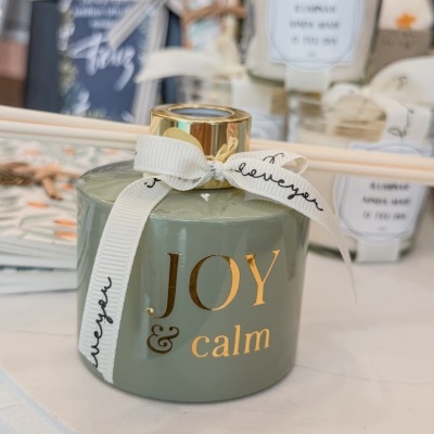 Frasco verde com tampa dourada e texto dourado 'JOY & calm' com fita branca amarrada