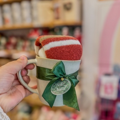Caneca branca com fita verde e etiqueta Feliz Natal com meia vermelha e branca içada à mão