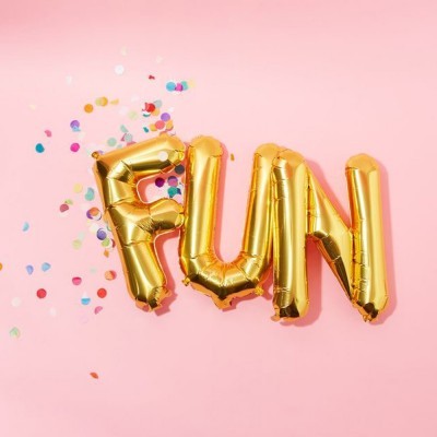Balões dourados formando a palavra FUN com confetes coloridos ao redor sobre fundo rosa