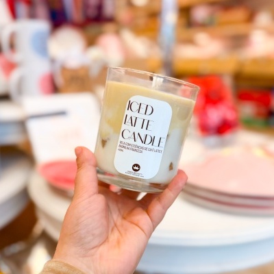 Vela de copo de vidro com etiqueta 'ICED LATTE CANDLE' segurada em mão, fundo desfocado com decoração doméstica.