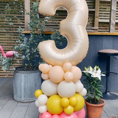 Balão dourado em forma do número 3 sobre balões menores coloridos, ao lado de plantas e parede de ripas de madeira