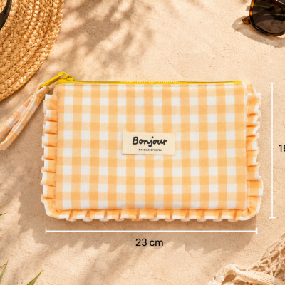 Necessaire de tecido xadrez amarelo e branco com fecho, junto de acessórios de verão sobre superfície bege.