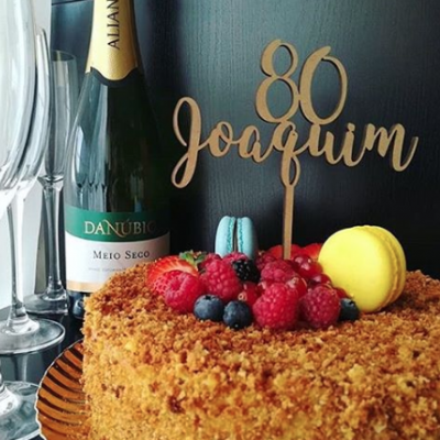 Bolo decorado com frutas e topper 80 Joaquim ao lado de garrafa de espumante e taças