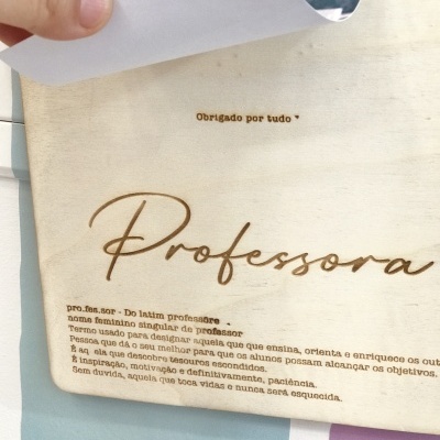 Folha de papel tipo pergaminho com texto sobre professora, segurada por uma mão, com parede colorida ao fundo.
