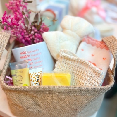 Cesto de presente com toalhas brancas, sabão amarelo, creme OLIVE OIL, caixa branca com texto vermelho, flores cor de rosa.