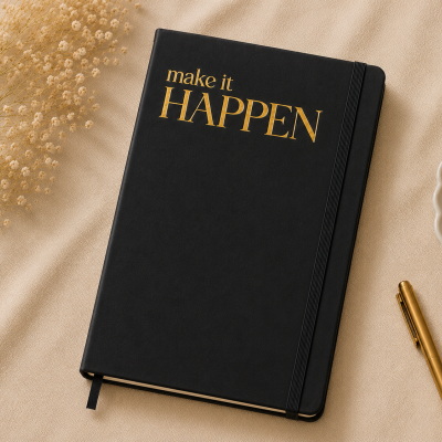 Caderno preto com texto dourado make it HAPPEN ao centro sobre tecido bege