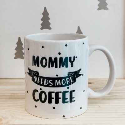 Caneca branca com texto 'MOMMY NEEDS MORE COFFEE' e pontos pretos num fundo claro.