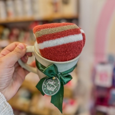 Caneca com laço verde e etiqueta 'Feliz Natal' contendo tecido colorido em tons vermelhos e bege