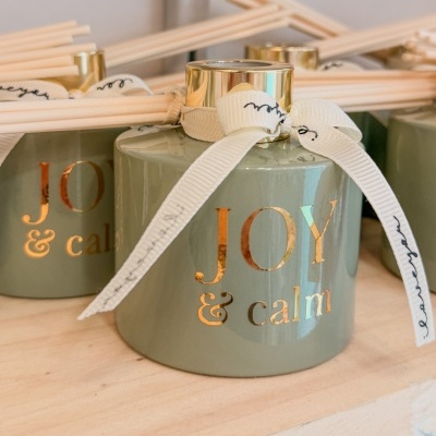 Difusores de ambiente verdes com texto JOY & calm e laços brancos na prateleira de madeira