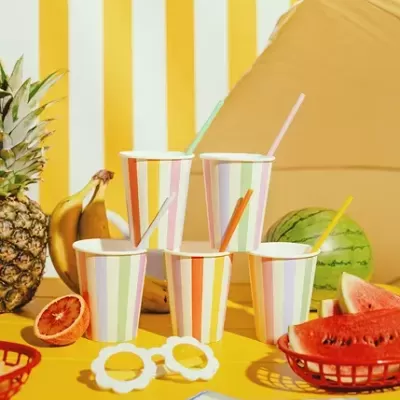 Cinco copos de papel coloridos, frutas e acessórios sobre mesa amarela
