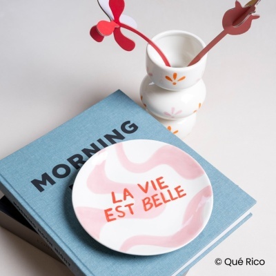 Prato branco com texto vermelho sobre livro azul e vaso branco decorado com marcadores vermelhos