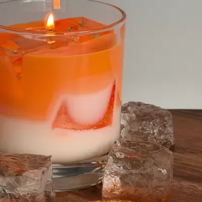 Vela laranja e branca acesa em copo com cubos de gelo e laranja seca