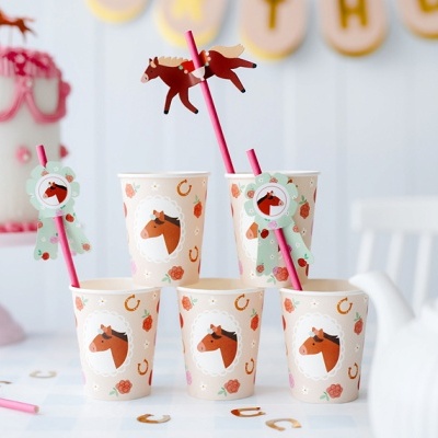 Copos de papel com desenhos de cavalos, ferraduras e flores, com palhinhas decoradas, em mesa branca perto de bolo decorado.