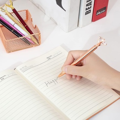 Mesa branca com caderno aberto e canetas decorativas e organizador de canetas cor cobre