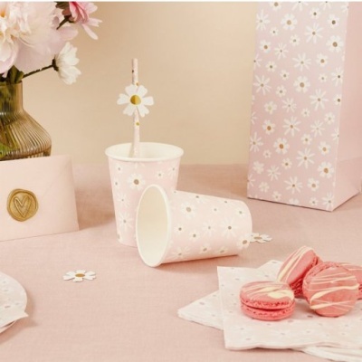 Copos e acessórios para festa em rosa claro com padrão de margaridas, mesa decorada com macarons e flores.