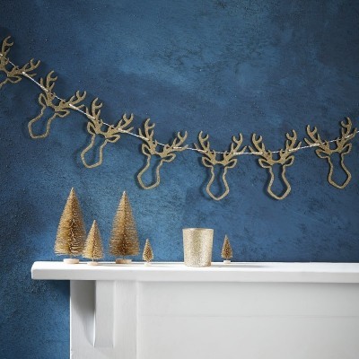 Corrente decorativa dourada com cabeças de veado numa parede azul acima de superfície branca com árvores e copo dourados