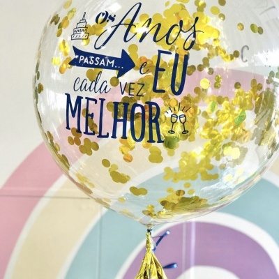 Balão transparente com confetes dourados e texto decorativo impresso.