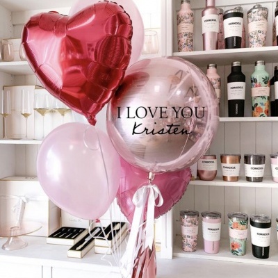 Balões decorativos cor de rosa com mensagem I LOVE YOU Kristen e garrafas decorativas em prateleiras ao fundo