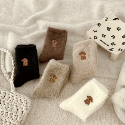 Meias felpudas com bordado de cão, em várias cores, sobre tecido branco com bolsa tricotada e banquinho decorado ao lado.