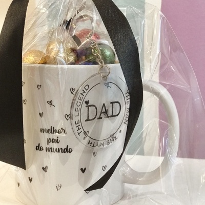 Caneca branca com texto e corações, embrulhada em plástico com fita preta e bombons coloridos