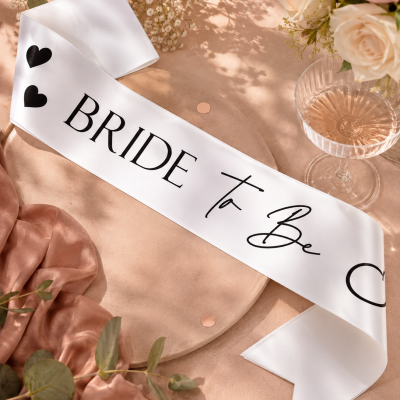 Faixa branca com texto Bride To Be em superfície decorada com flores, tecido rosa e confetes.