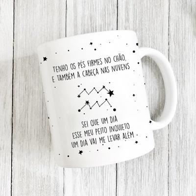 Caneca branca com texto em português e desenho de constelação sobre mesa de madeira clara.