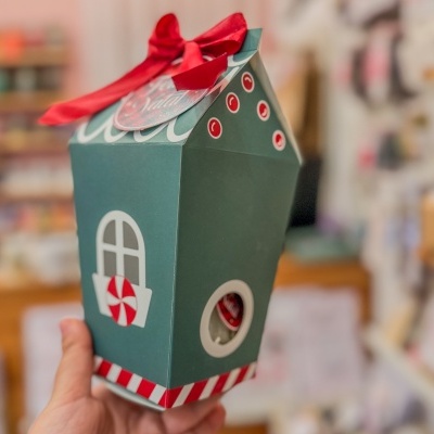 Embalagem natalícia em forma de casa verde com decoração vermelha e branca sendo segurada