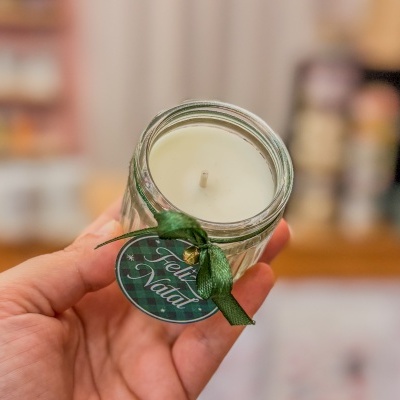 Vela branca em frasco de vidro com etiqueta 'Feliz Natal' e fita verde.