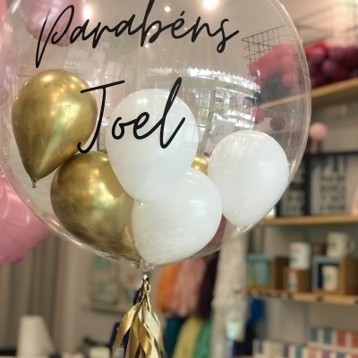 Balão transparente com texto 'Parabéns Joel' e balões internos dourados e brancos