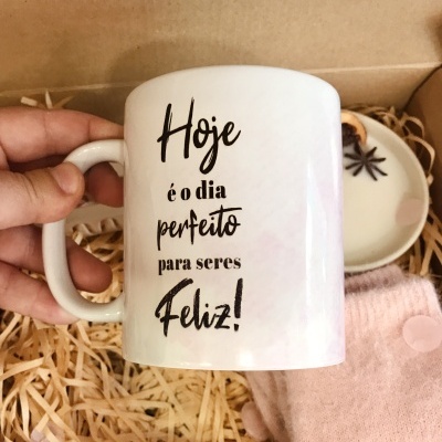 Caneca branca com frase em português sobre fundo com palha de embalar, prato branco e tecido rosa.