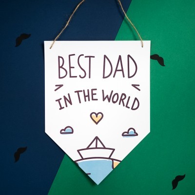 Pendurante branco com texto 'BEST DAD IN THE WORLD' e ilustrações coloridas, pendurado por cordão rústico.