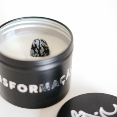 Vela branca em recipiente preto com tampa preta decorada com desenho branco de mão e palavra TRANSFORMAÇÃO no recipiente.