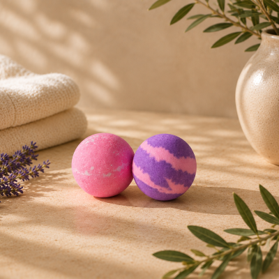 Bolas de banho rosa e roxa com toalha creme, lavanda e vaso cerâmico em ambiente iluminado