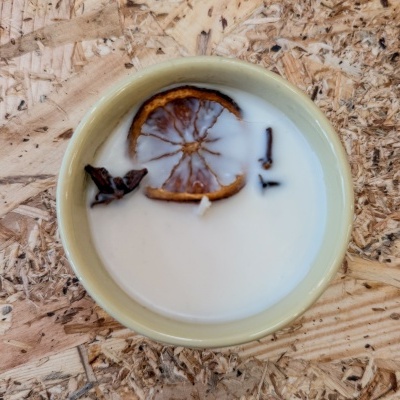 Vela branca num recipiente cerâmico com rodela de laranja seca e cravinhos sobre madeira clara