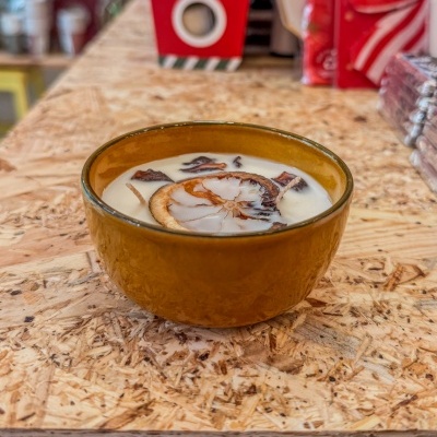 Vela decorativa branca num recipiente de cerâmica castanha com pedaços de casca ou flores secas.