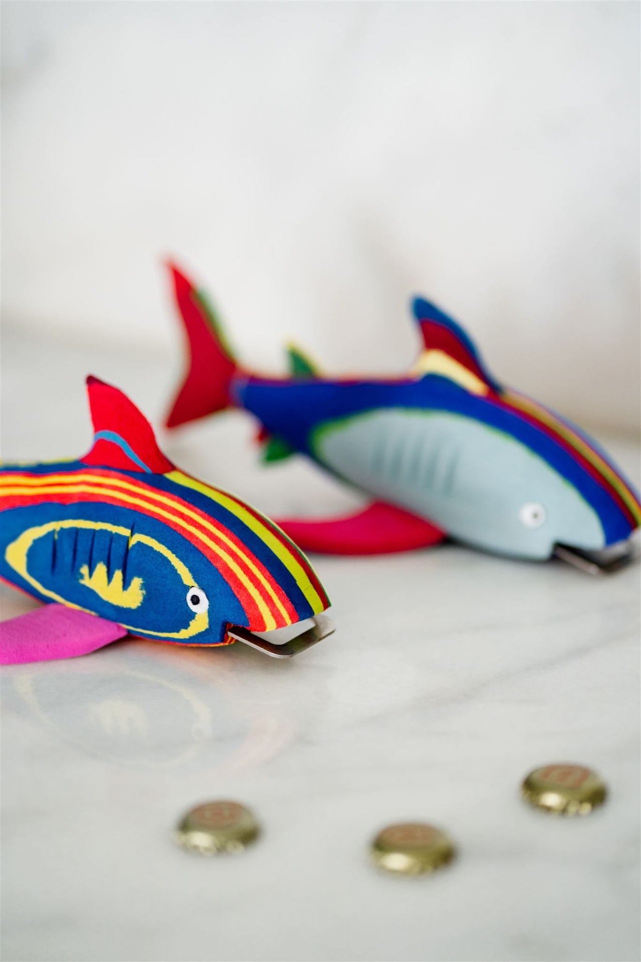 Tubarão Abre Garrafas Dois peixes de tecido colorido com padrões listrados em azul, vermelho, amarelo e rosa sobre uma superfície branca