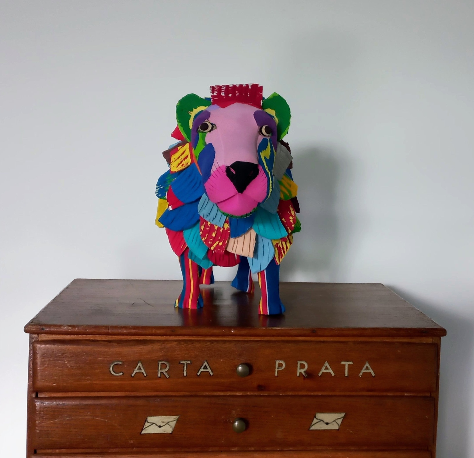 Leão L Estatueta colorida de animal em tecido sobre móvel de madeira com inscrição CARTA PRATA