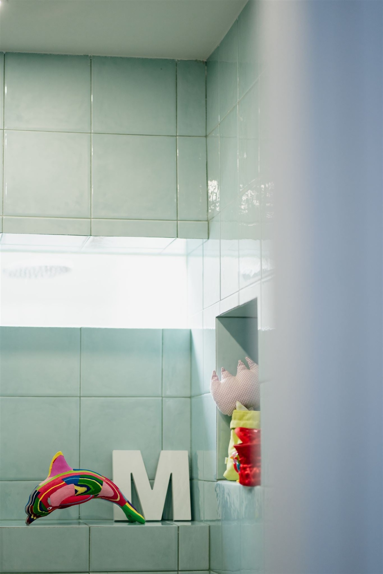Golfinho M Paredes com azulejos verdes claros, janela, letra M branca, almofada cor de rosa, brinquedo colorido