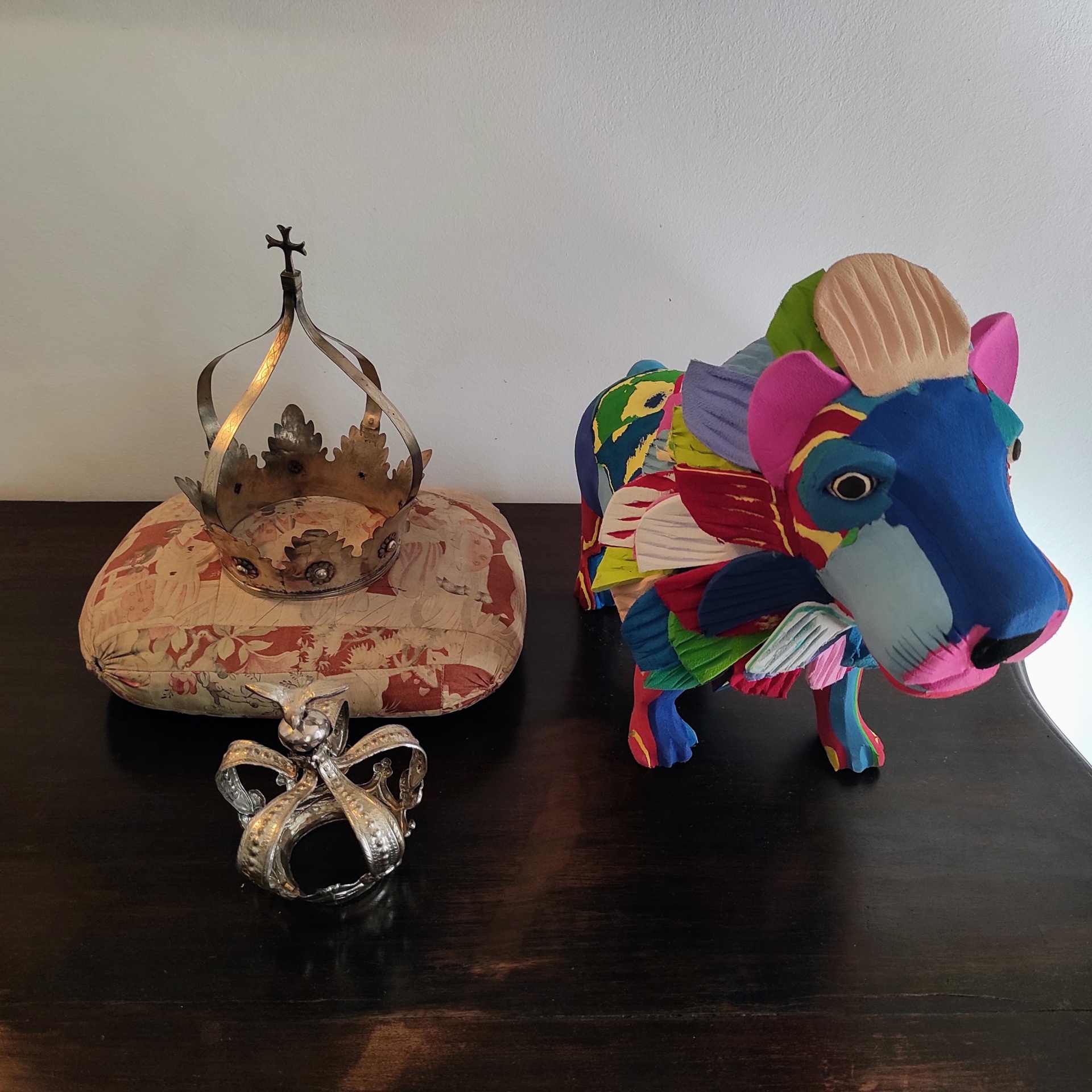 Leão L Estatueta colorida de animal em madeira ao lado de duas coroas em cima de balcão escuro.