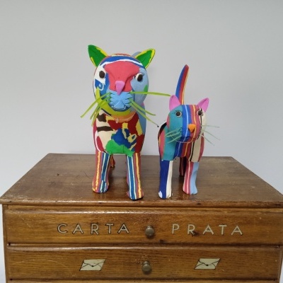 Duas esculturas de gatos coloridos em tecido sobre uma gaveteira de madeira com inscrições.