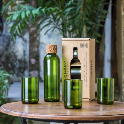 garrafa verde com tampa de madeira e três copos verdes sobre mesa redonda em ambiente exterior