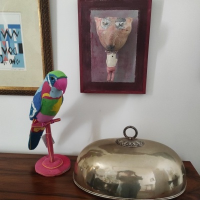 Papagaio de madeira colorido e peça metálica sobre mesa de madeira com quadros na parede branca