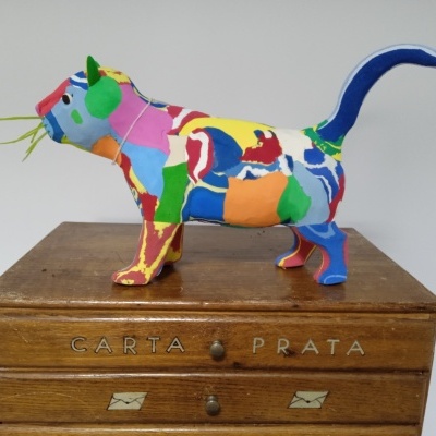 Estatueta colorida de gato sobre móvel de madeira com inscrições.