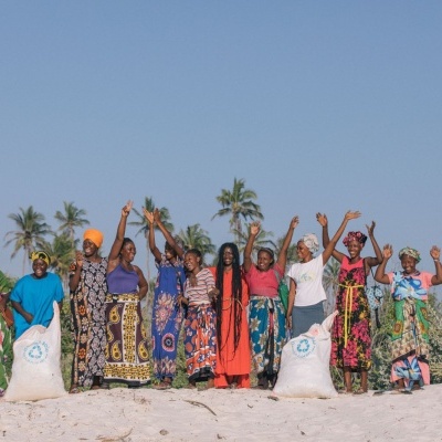 Dez mulheres africanas vestidas com roupas coloridas em praia com sacas brancas e palmeiras ao fundo