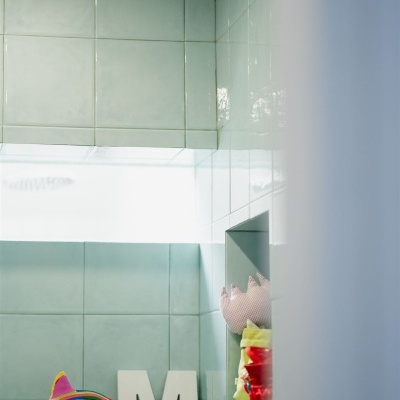 Paredes com azulejos verdes claros, janela, letra M branca, almofada cor de rosa, brinquedo colorido