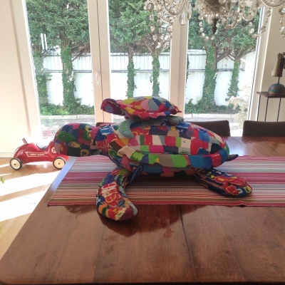 Almofada em forma de tartaruga colorida sobre mesa de madeira com manta riscada frente a janela