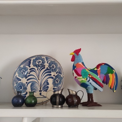 Decoração numa prateleira com prato cerâmico azul e branco, potes pequenos coloridos, jarras em vidro e escultura de galo de tecido colorido.